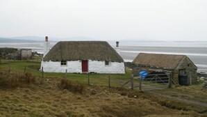 Une maison traditionnelle à toit de chaume, avec une porte rouge, située près d’une plage et offrant une vue sur la mer et les collines lointaines.