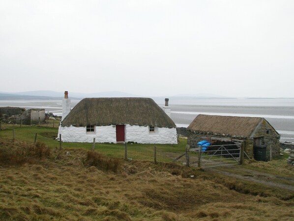Una casa tradicional con techo de paja y puerta roja, ubicada cerca de una playa con vista al mar y colinas lejanas.