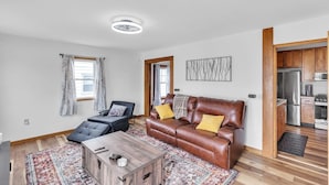 Una sala moderna con un sofá de cuero marrón, un sillón negro y una mesa de centro de madera. Se ve una cocina con armarios de madera y electrodomésticos de acero inoxidable a través de un vano abierto.
