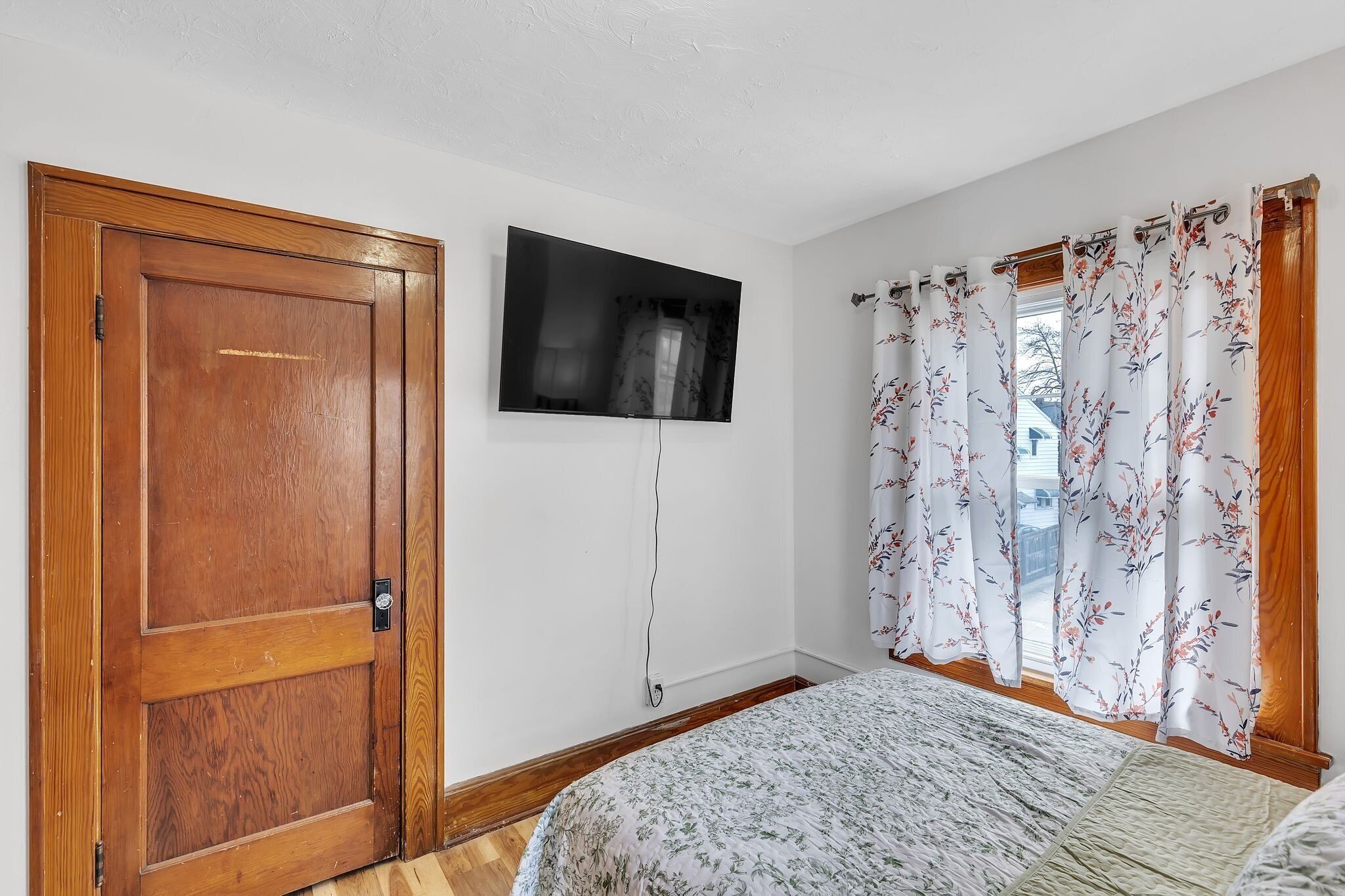 Une chambre avec une porte en bois, un téléviseur à écran plat fixé au mur, des rideaux à motifs floraux et un lit recouvert d’une couvre-lit à motifs.