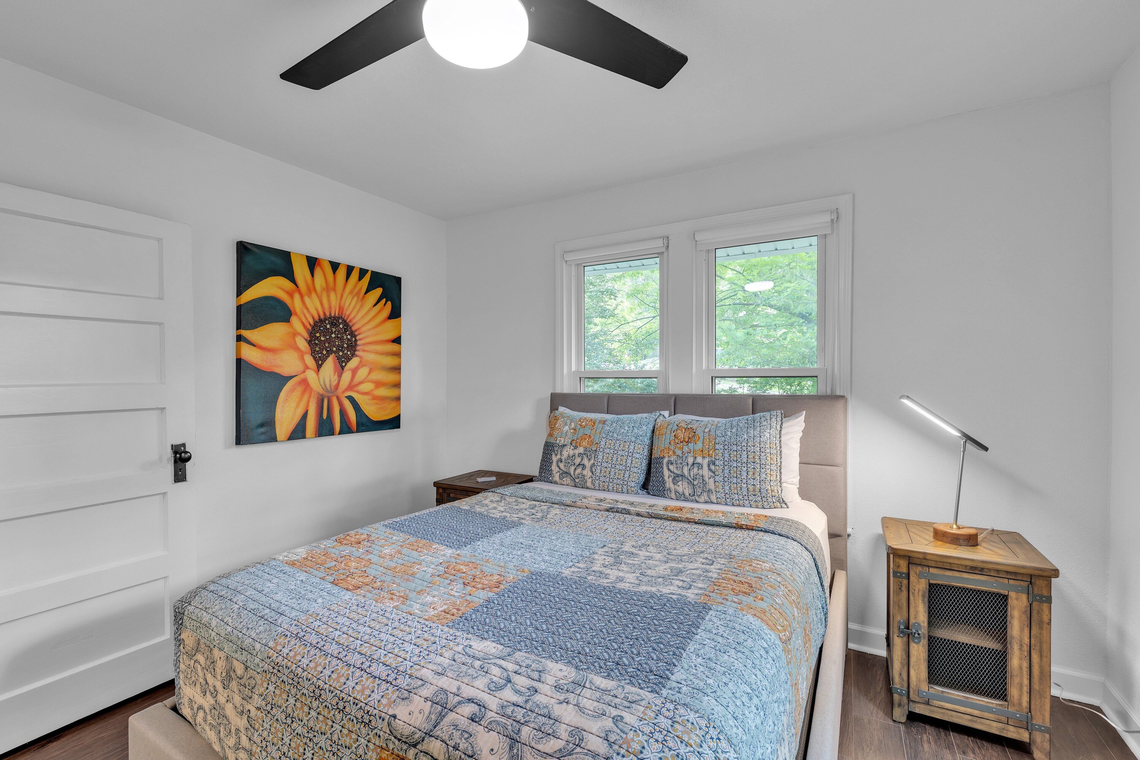 A bedroom with a large bed, a ceiling fan, a window with a view of greenery, and a nightstand with a lamp.