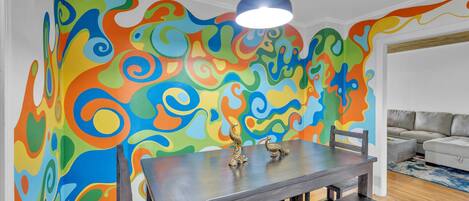 Uma área de refeições com um mural abstrato e colorido, uma mesa moderna e uma luminária contemporânea.