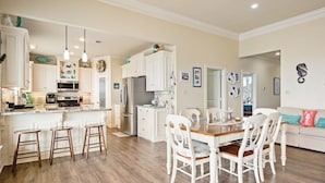 A spacious kitchen with white cabinetry, a central island with stools, and a dining area with white chairs. - Pelican Path 3 Bedroom Home (Port Bolivar)