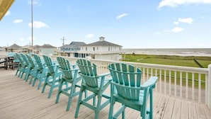 A deck with turquoise Adirondack chairs, a white railing, and a view of the ocean and beach houses. - Pelican Path 3 Bedroom Home (Port Bolivar)