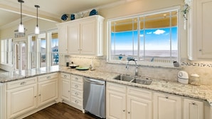 A kitchen with white cabinets, granite countertops, a dishwasher, and a large window with a view of the ocean. - Pelican Path 3 Bedroom Home (Port Bolivar)