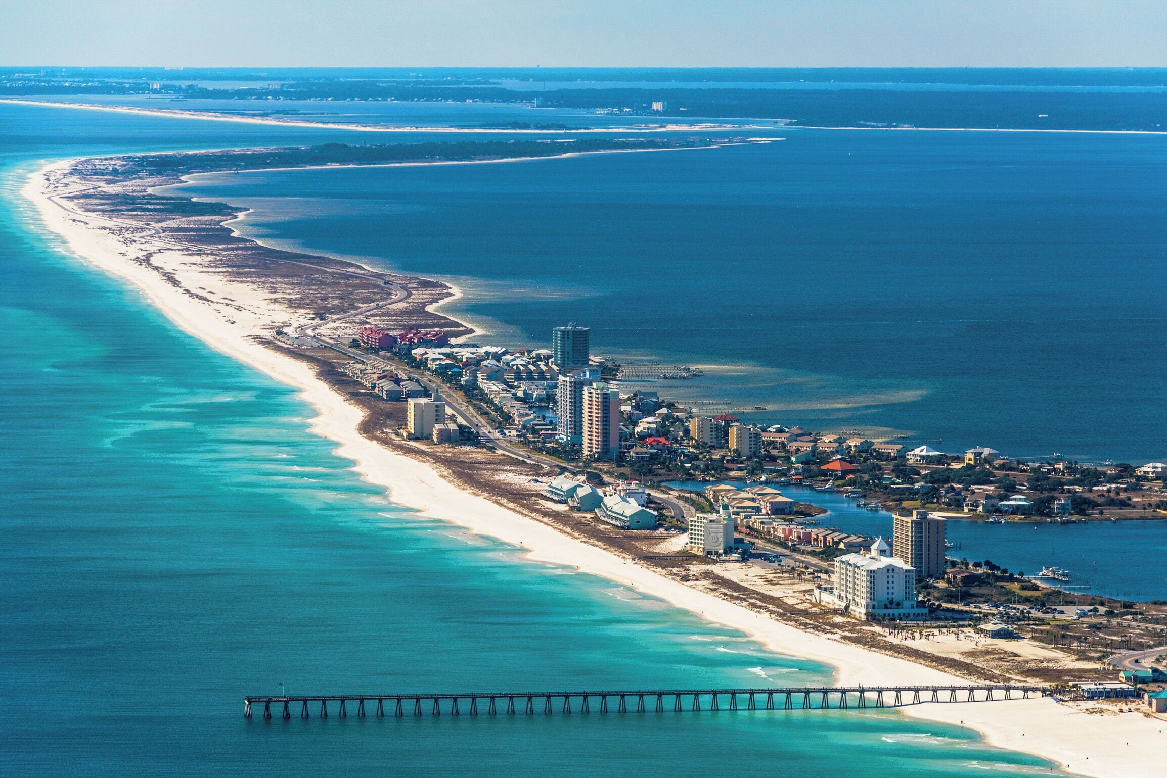 An aerial view of a coastal area with a long beach, a pier, and a cluster of buildings.