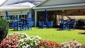 Ein überdachter Außenbereich mit blauen Stühlen und Sonnenschirmen, eine Blumenwiese und ein Gebäude mit weißer Fassade und Säulen.