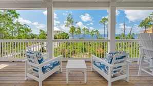 Un espace extérieur aménagé, abrité et doté de chaises blanches et d’une table, offrant une vue sur une plage bordée d’arbres et un ciel dégagé.