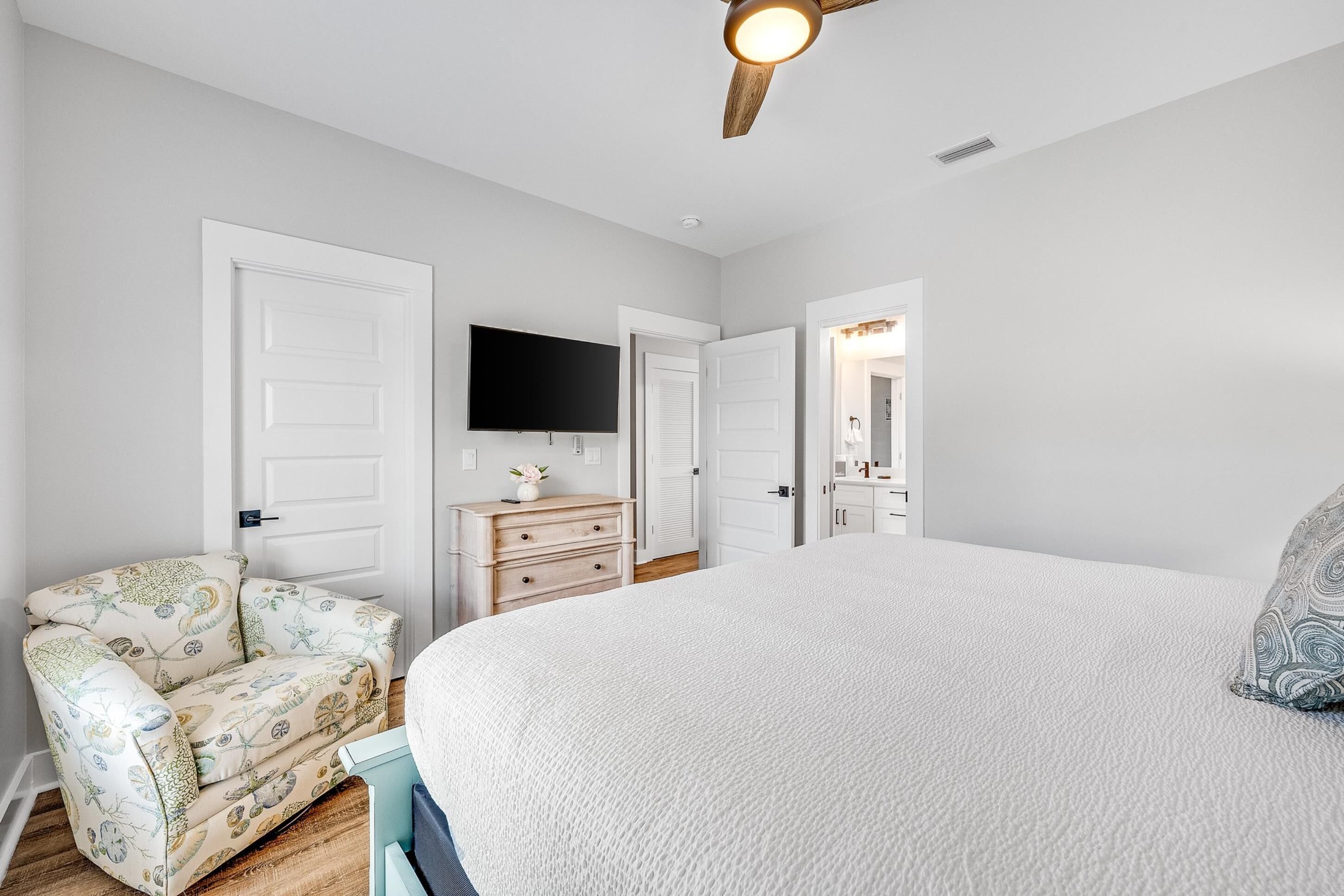 Une chambre avec un grand lit, un ventilateur de plafond, un téléviseur fixé au mur et une commode avec des tiroirs.