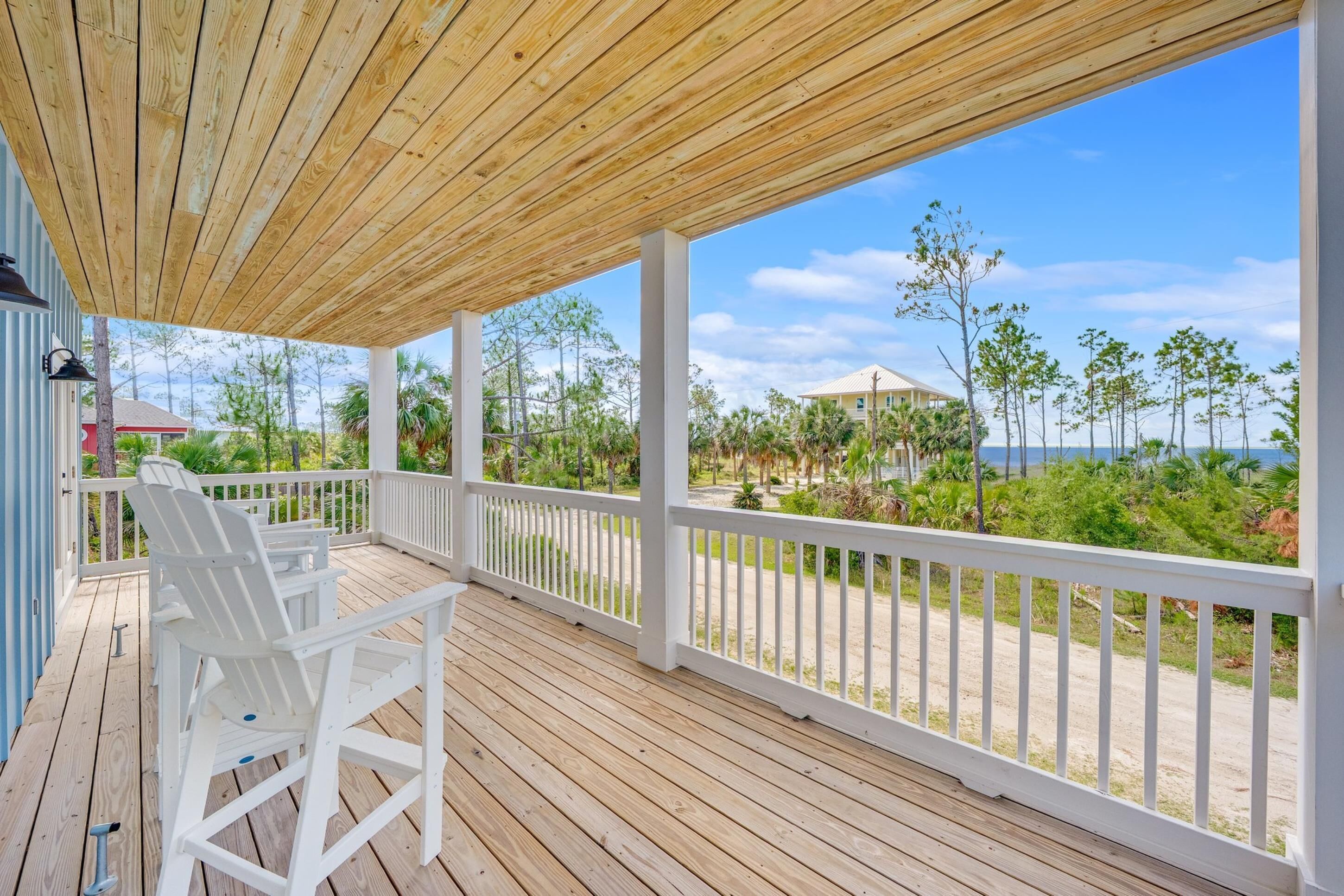 A wooden deck with white railings and chairs, overlooking a beach and buildings.