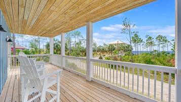 Una terraza de madera con barandas blancas y sillas, con vista a una playa y edificios.
