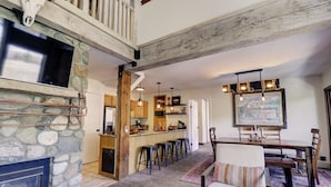 A spacious living area with a stone fireplace, a dining table with chairs, and a kitchen in the background.