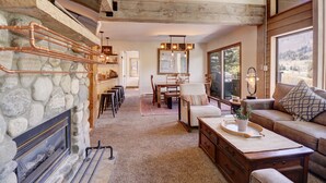 A cozy living room with a stone fireplace, wooden beams, a dining area, and a large window offering a view of the outdoors.