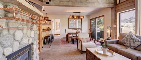 Una sala de estar acogedora con chimenea de piedra, vigas de madera, comedor y un amplio ventanal con vistas al exterior.