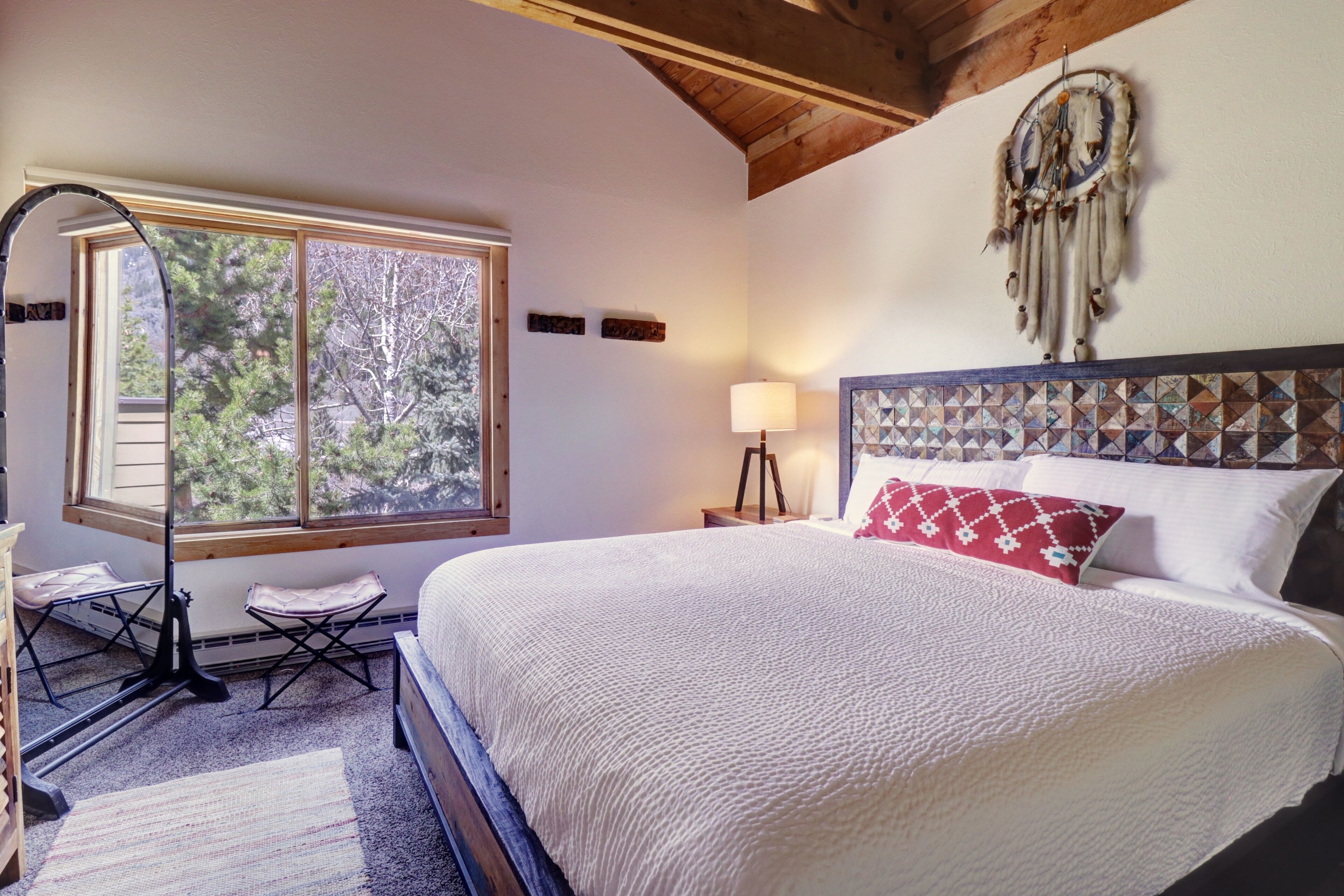 A bedroom with a large bed, a window with a view of trees, a lamp, and a dreamcatcher on the wall.