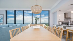 A modern dining area with a large wooden table, a bowl of fruit, and a view of the ocean through large windows. - Phoenix Gulf Shores Ii 2201 4 Bedroom Condo (Gulf Shores)
