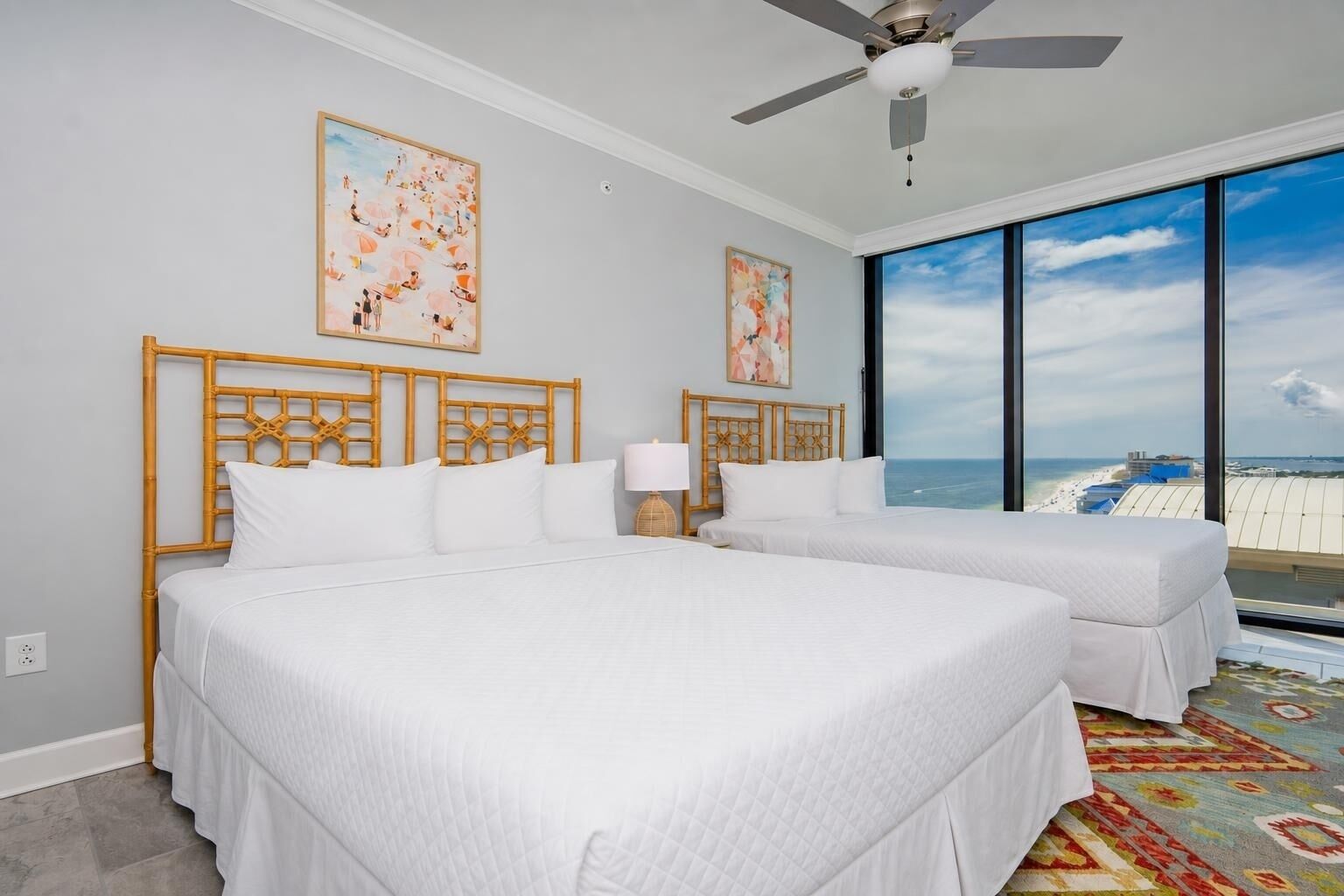 Habitación de hotel con dos camas, ventilador de techo, ventanas amplias con vista a la playa y obras de arte en las paredes.