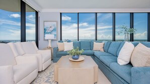 A modern living room with a large window overlooking the sea, a blue sofa, a white armchair, and a coffee table with a plant. - Phoenix Gulf Shores Ii 2201 4 Bedroom Condo (Gulf Shores)