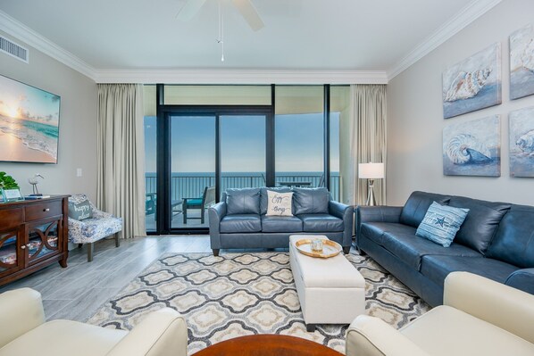 A modern living room with a grey leather sofa, a white ottoman, and a patterned rug. A wooden side table with a lamp is visible. - Phoenix Gulf Shores Ii 2404 3 Bedroom Condo (Gulf Shores)
