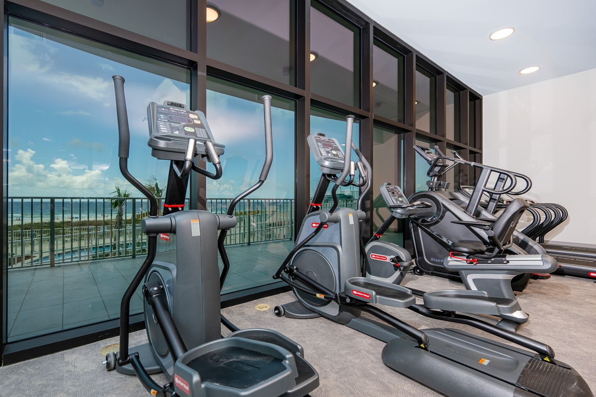 Filas de bicicletas estáticas y cintas de correr en un gimnasio con amplias ventanas con vistas a la playa.