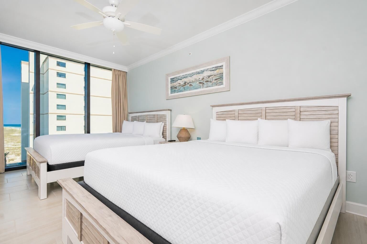 Habitación de hotel con dos camas, ventilador de techo, un cuadro y vista al océano.
