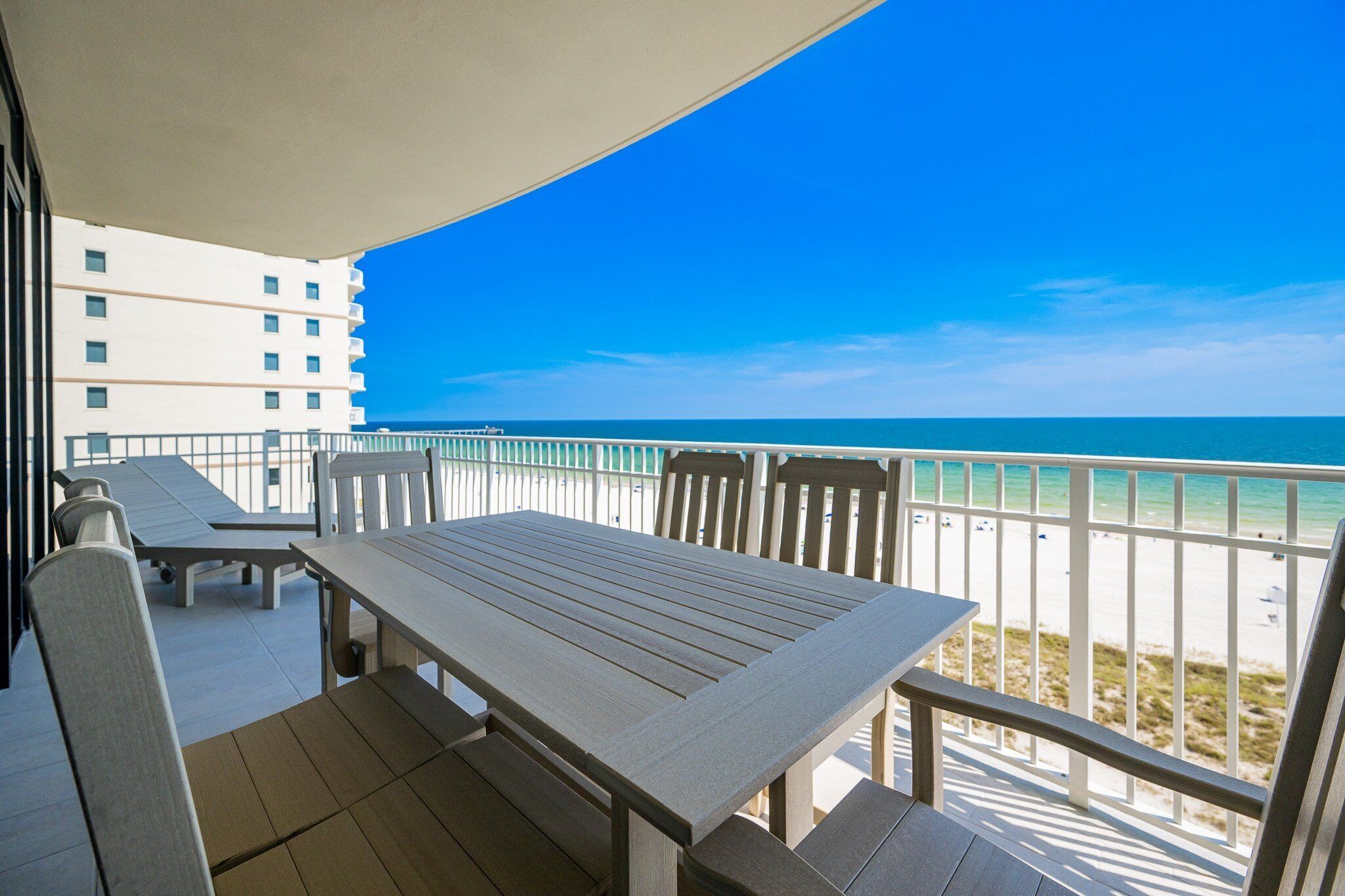 A balcony with a table and chairs, overlooking a beach with a clear blue sky and ocean.