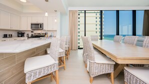 Cozinha moderna com armários brancos, micro-ondas e máquina de café. Há uma área de refeições com cadeiras de vime e uma mesa de madeira. O ambiente possui grandes janelas com vista para edifícios e um corpo d'água.