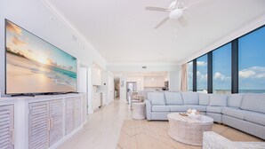 Uma sala de estar ampla com uma TV de tela plana grande, um sofá seccional cinza e vista para o oceano através de grandes janelas.