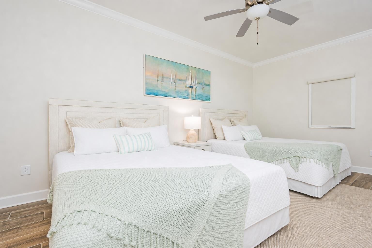 Une chambre d’hôtel avec deux lits, un ventilateur de plafond, un tableau représentant des voiliers et une photo encadrée accrochée au mur.