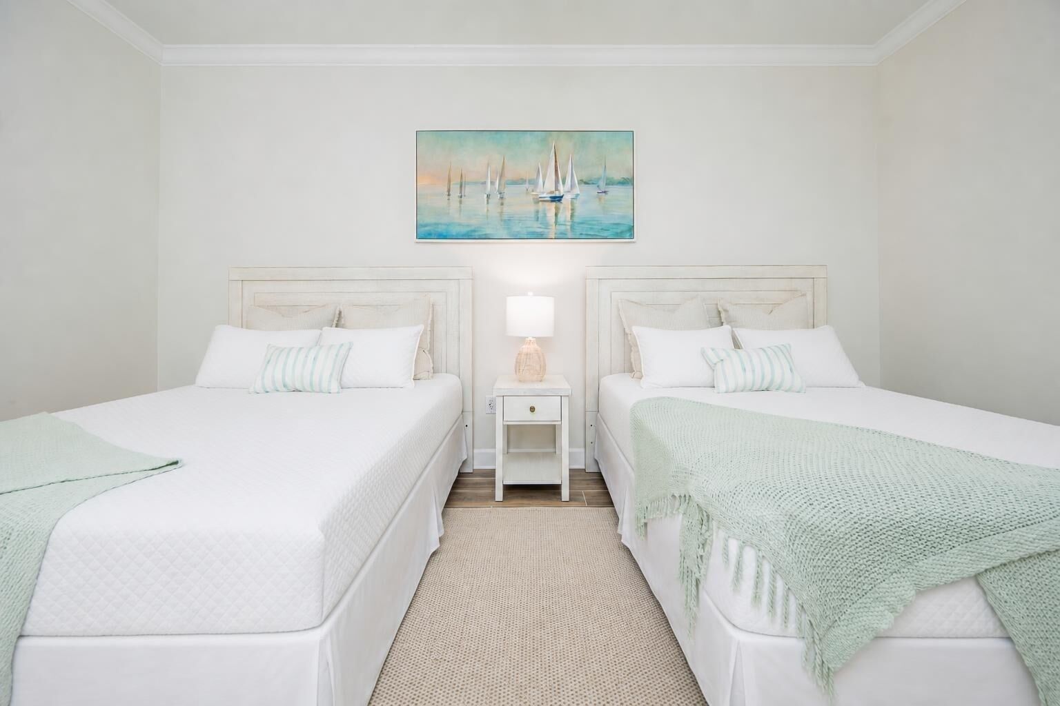 A hotel room with two beds, a nightstand, a lamp, and a painting of sailboats.