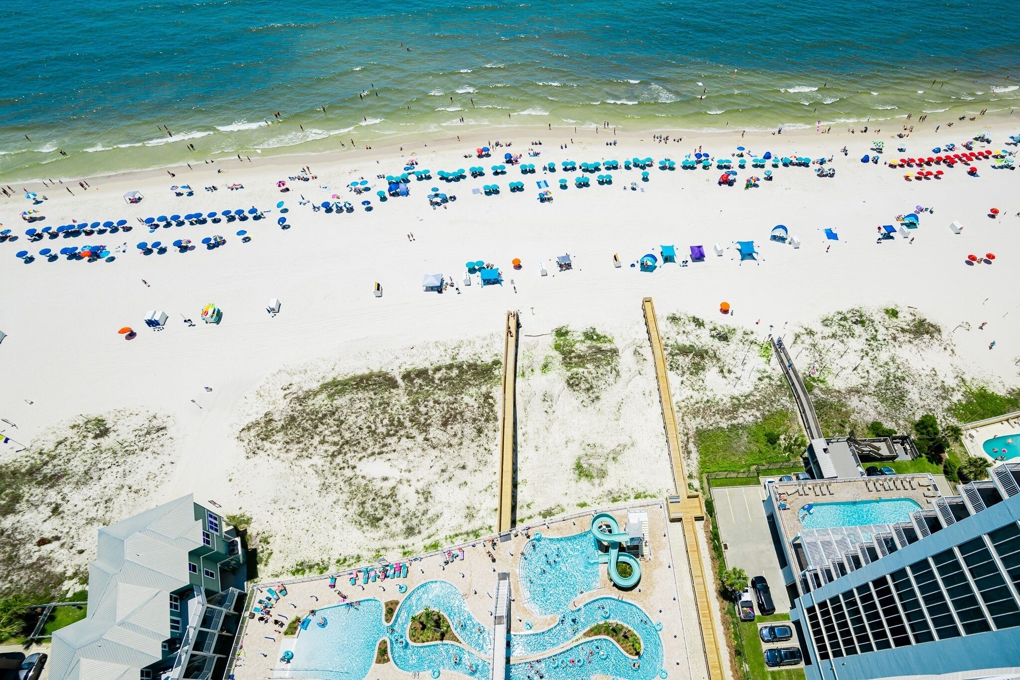 A beach with numerous umbrellas and people, a pool area with multiple swimming pools, and buildings with balconies overlooking the beach.
