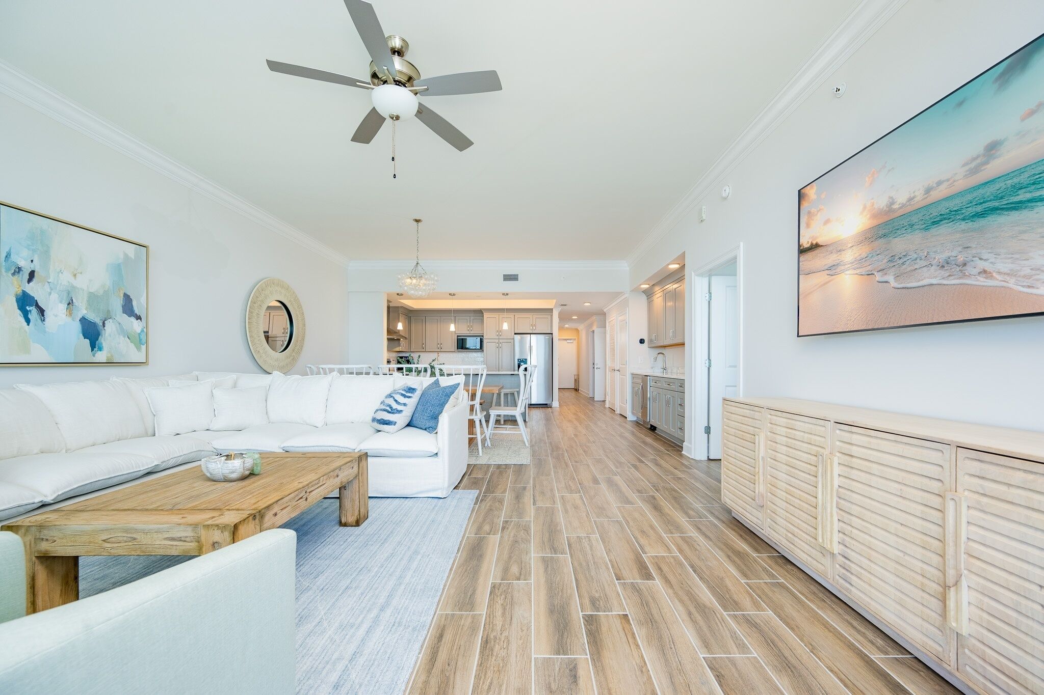A spacious living area with a white sofa, a wooden coffee table, and a ceiling fan. A kitchen is visible in the background with white cabinets and a large window.