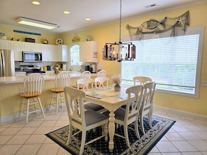 A dining area with a table set for six, surrounded by white chairs. A chandelier hangs above, and a window with blinds is visible.