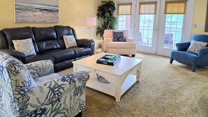 A living room with a black leather sofa, a white armchair, a blue armchair, and a coffee table. There is a painting on the wall and a plant in the corner. - Peaceful 3BR Ground Floor Condo w/ Intracoastal Waterway Views! (Myrtle Beach)