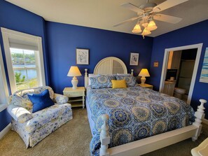 A bedroom with a large bed, two bedside tables, a chair, a ceiling fan, and a window with a view of a body of water.