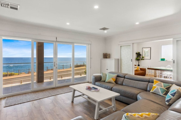 A modern, stylish living room with a large sectional sofa adorned with colorful cushions. The room features expansive sliding glass doors that offer a stunning view of the ocean, creating a serene and inviting atmosphere. The interior is bright and airy,