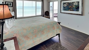 A bedroom with a large bed, a view of the ocean through the window, and a framed painting on the wall.