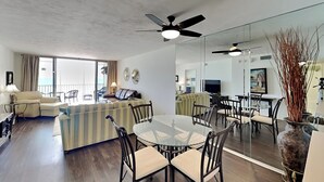 A modern living room with a glass dining table, dark wood flooring, and a large mirror reflecting the space.