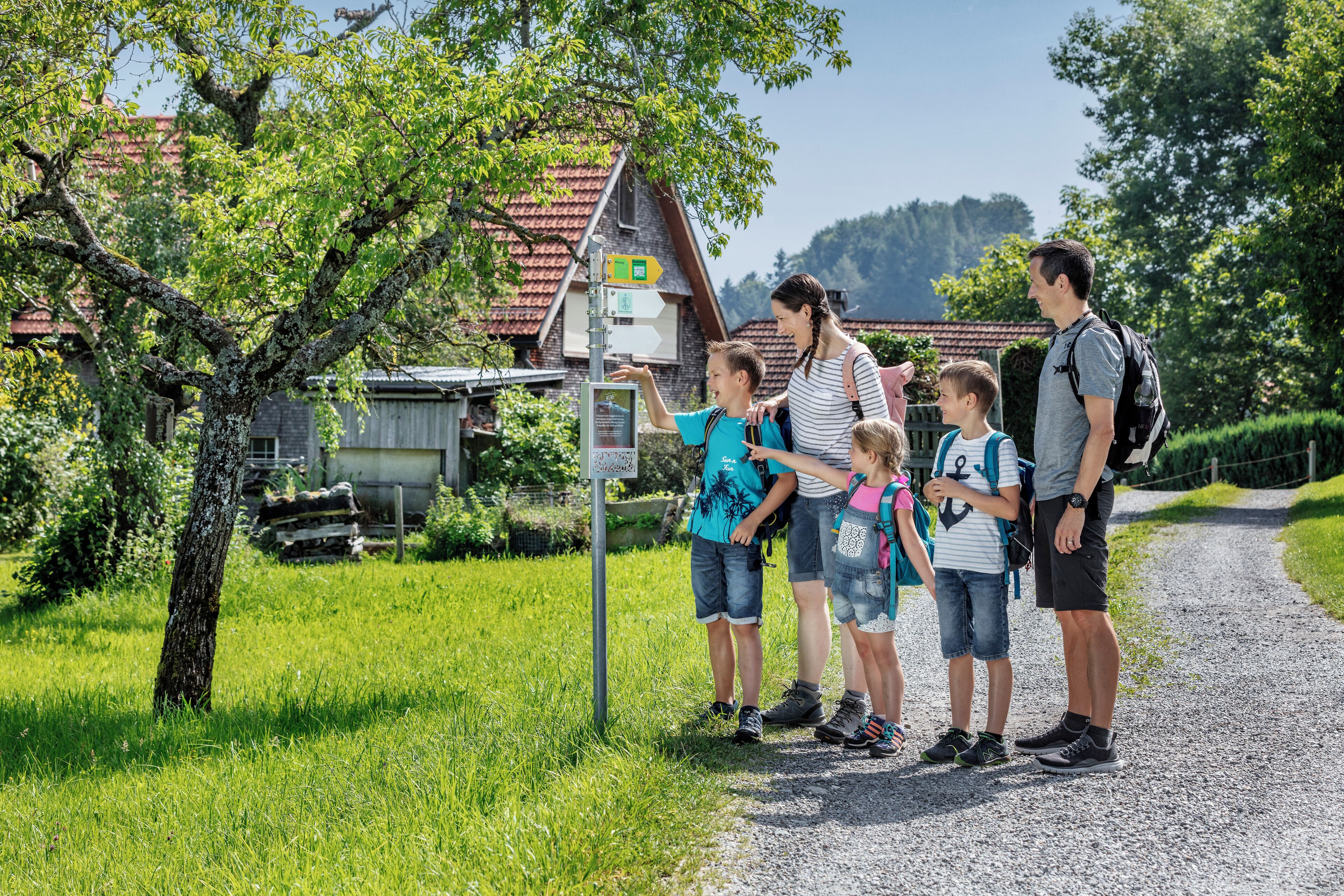 Eine Familie steht auf einem Schotterweg in ländlicher Umgebung, ein Wegweiser ist zu sehen.