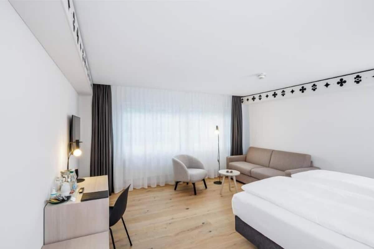 Moderni hotellihuone, jossa on puulattia, sänky, työpöytä tuolilla, sohva ja pieni pöytä.