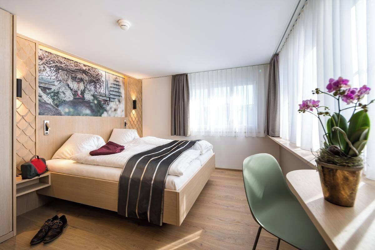Moderni hotellihuone, jossa on sänky, työpöytä, tuoli, ikkuna verhoilla ja seinälle kiinnitetty taideteos.