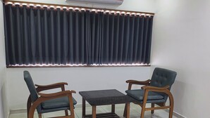 Une pièce comprenant deux chaises en bois, une petite table, un ventilateur de plafond, un appareil de climatisation et une grande fenêtre avec des rideaux foncés.
