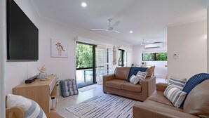 A modern living room with a sofa, a coffee table, a TV, and a ceiling fan. - Liana One Coastal Mission Vibe (Mission Beach)