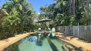 A clear swimming pool surrounded by lush tropical vegetation, with a fence and a shaded area with chairs. - Liana One Coastal Mission Vibe (Mission Beach)