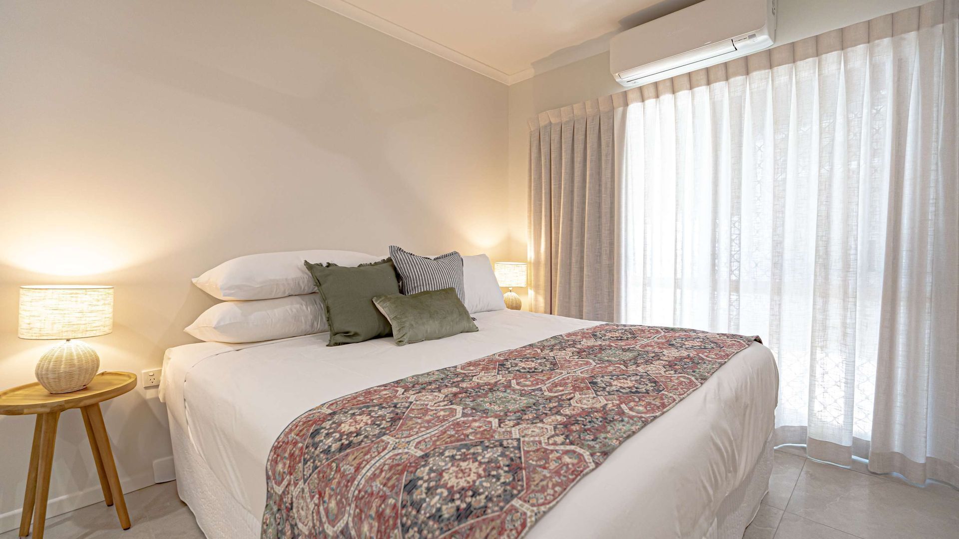 A bedroom with a bed, pillows, a bedside lamp, a small table, and a window with curtains.