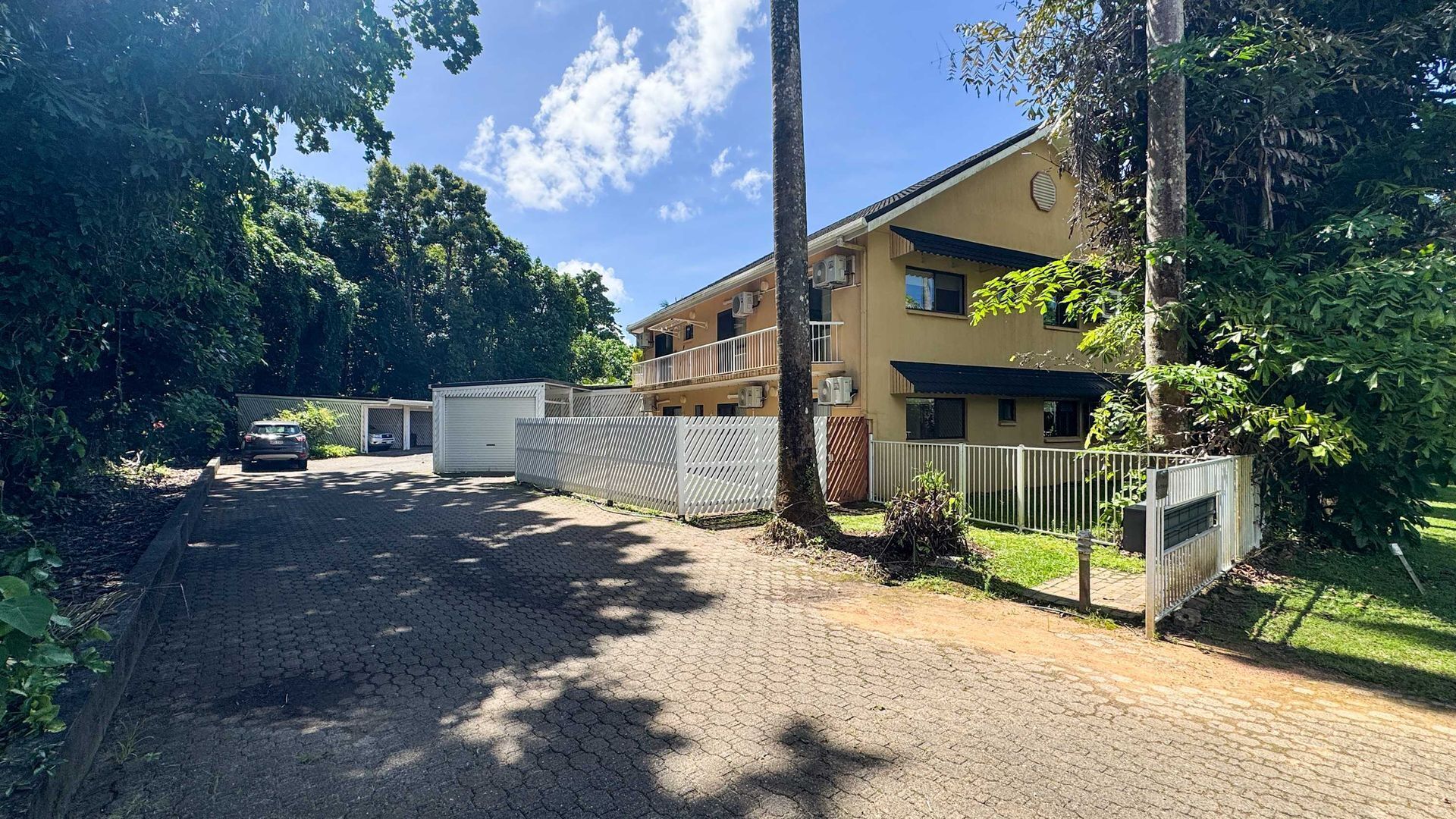 A residential building with a paved driveway, a fenced parking area, and lush greenery.