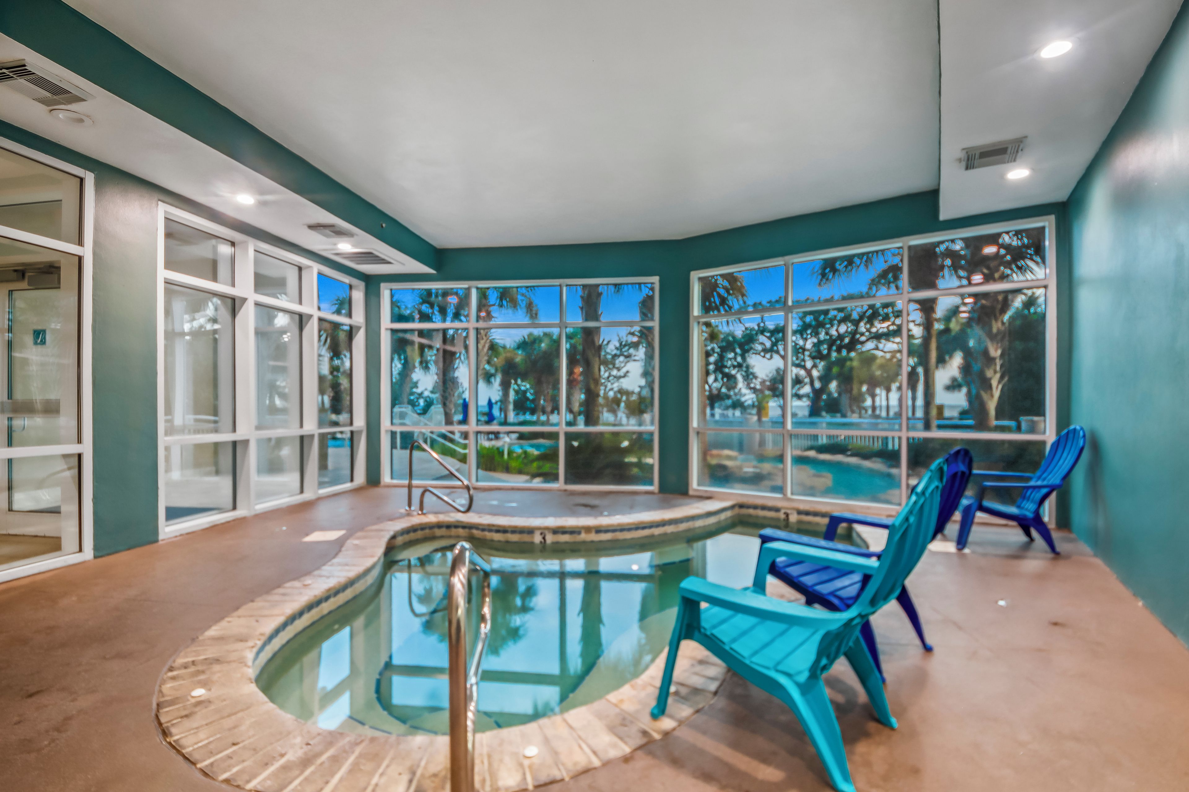 An indoor swimming pool with clear glass walls, blue chairs, and a view of palm trees outside.