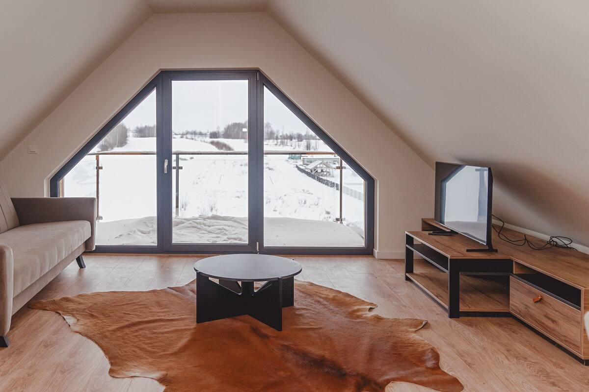 A modern loft bedroom with a sloped ceiling, a large sliding glass door leading to a snowy outdoor area, a leather sofa, a coffee table, and a TV on a wooden stand.