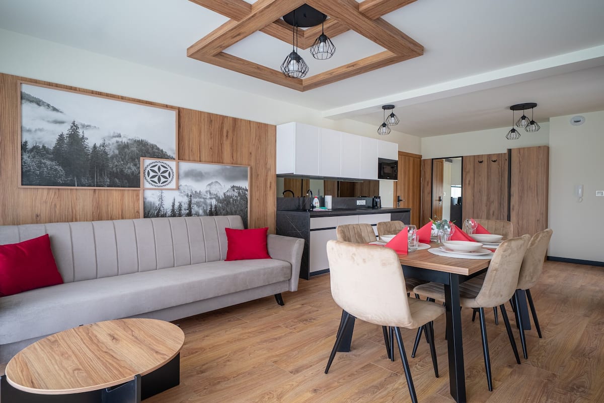 A modern living room with a grey sofa, red cushions, a wooden coffee table, and a dining area with a wooden table and chairs.