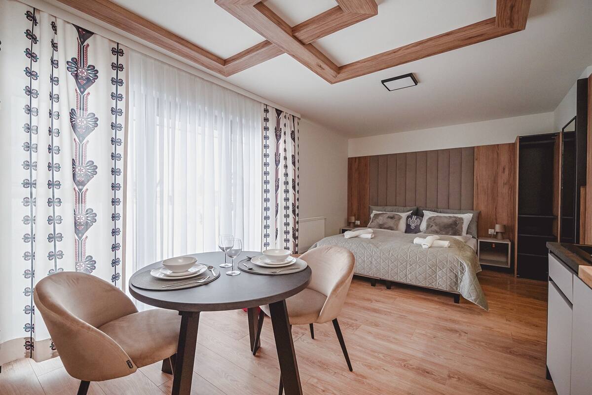 A modern bedroom with a wooden ceiling, a bed with pillows, a bedside table, and a window with patterned curtains.